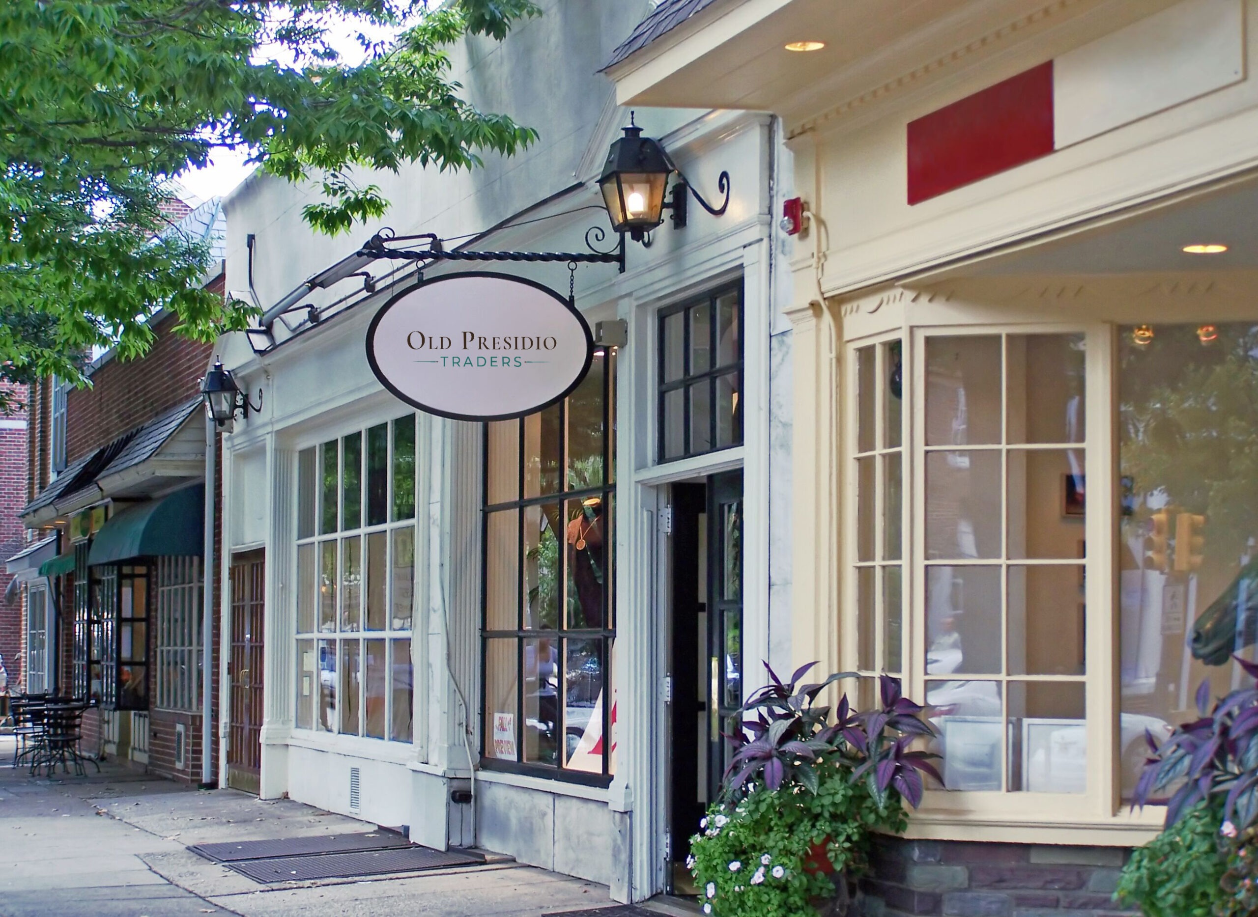 A quaint street scene featuring a shopfront with large windows and a sign reading "Old Presidio Traders." Hanging lanterns and potted plants add charm.