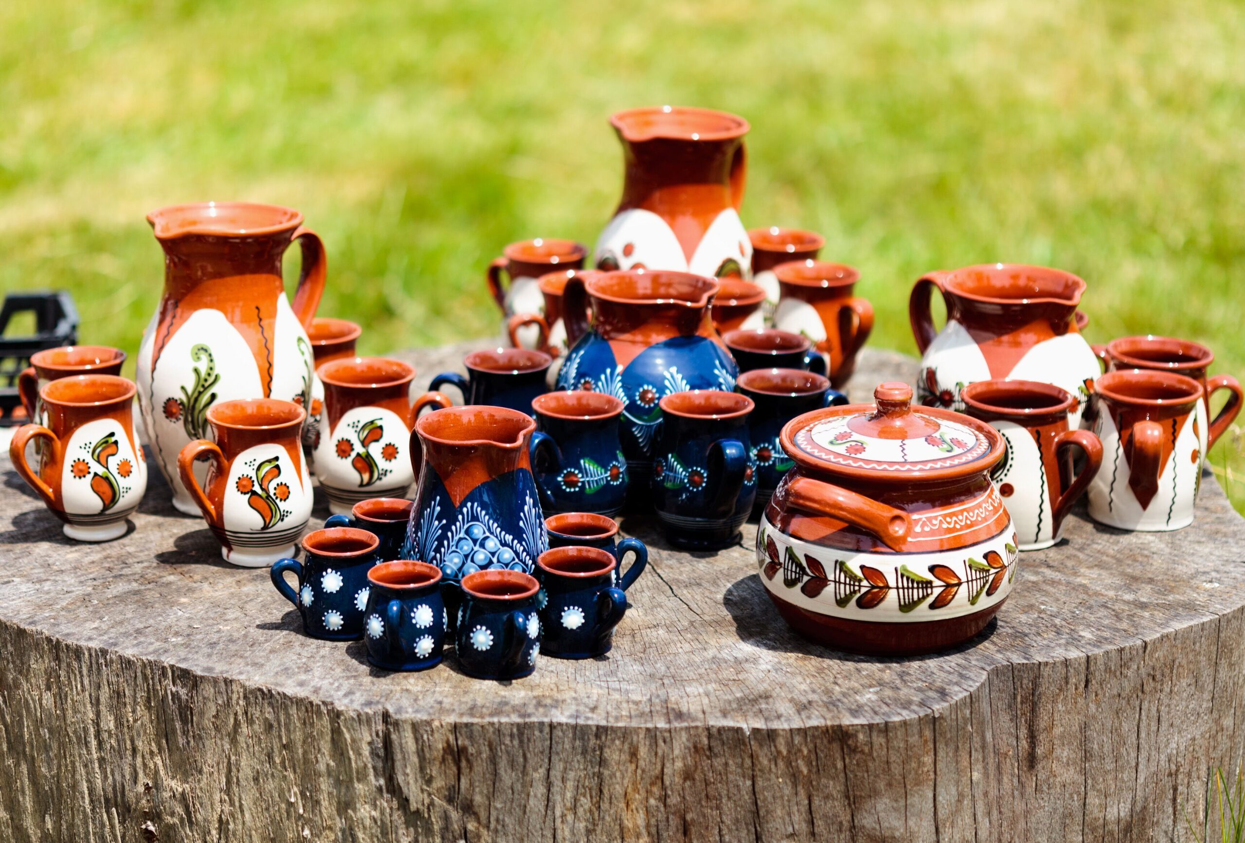 A colorful assortment of hand-painted pottery, including blue and white mugs, pitchers, and a lidded jar, displayed on a wooden stump in a green grassy area.