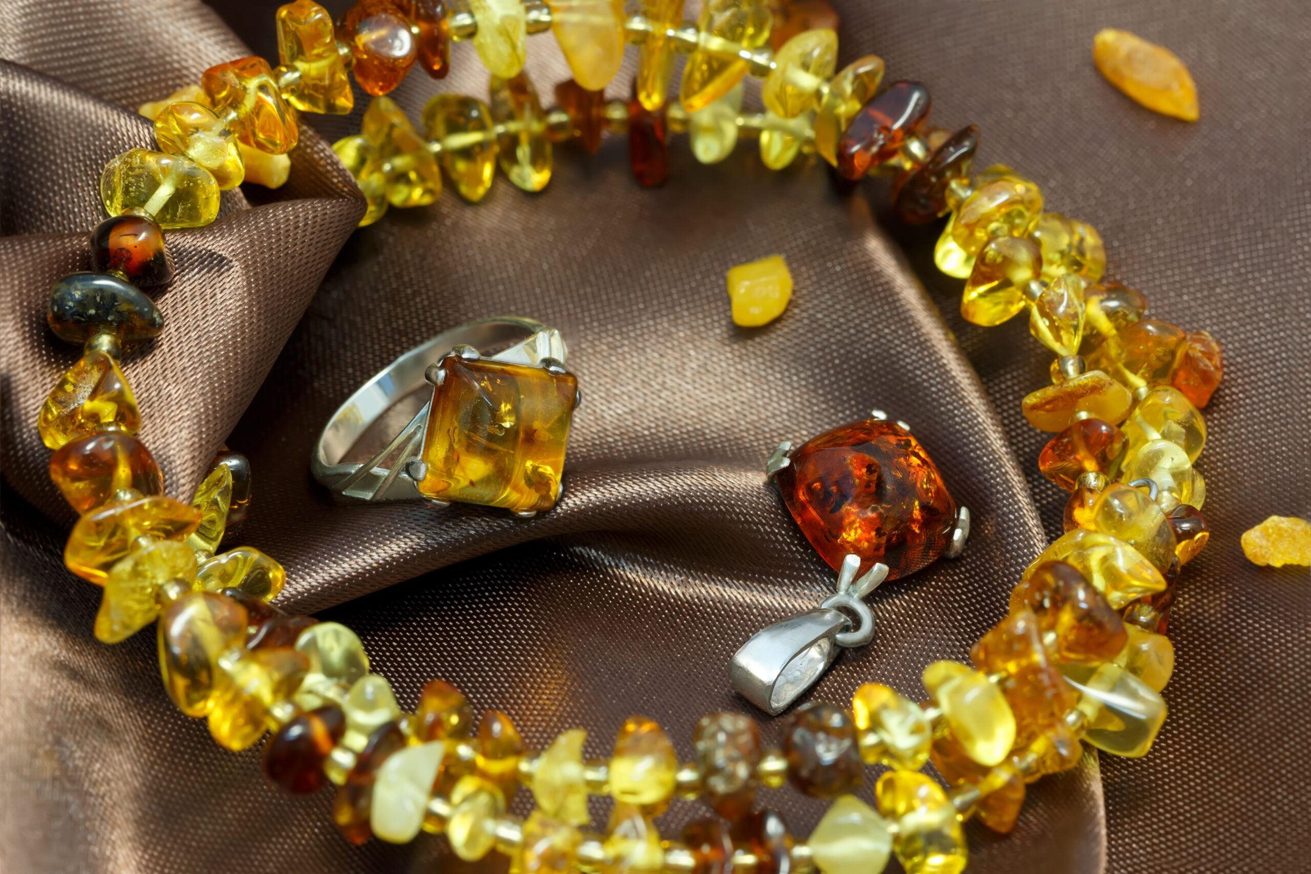 Amber jewelry on brown satin: a bracelet of multicolored amber stones, an amber ring, and a pendant, conveying elegance and warmth.