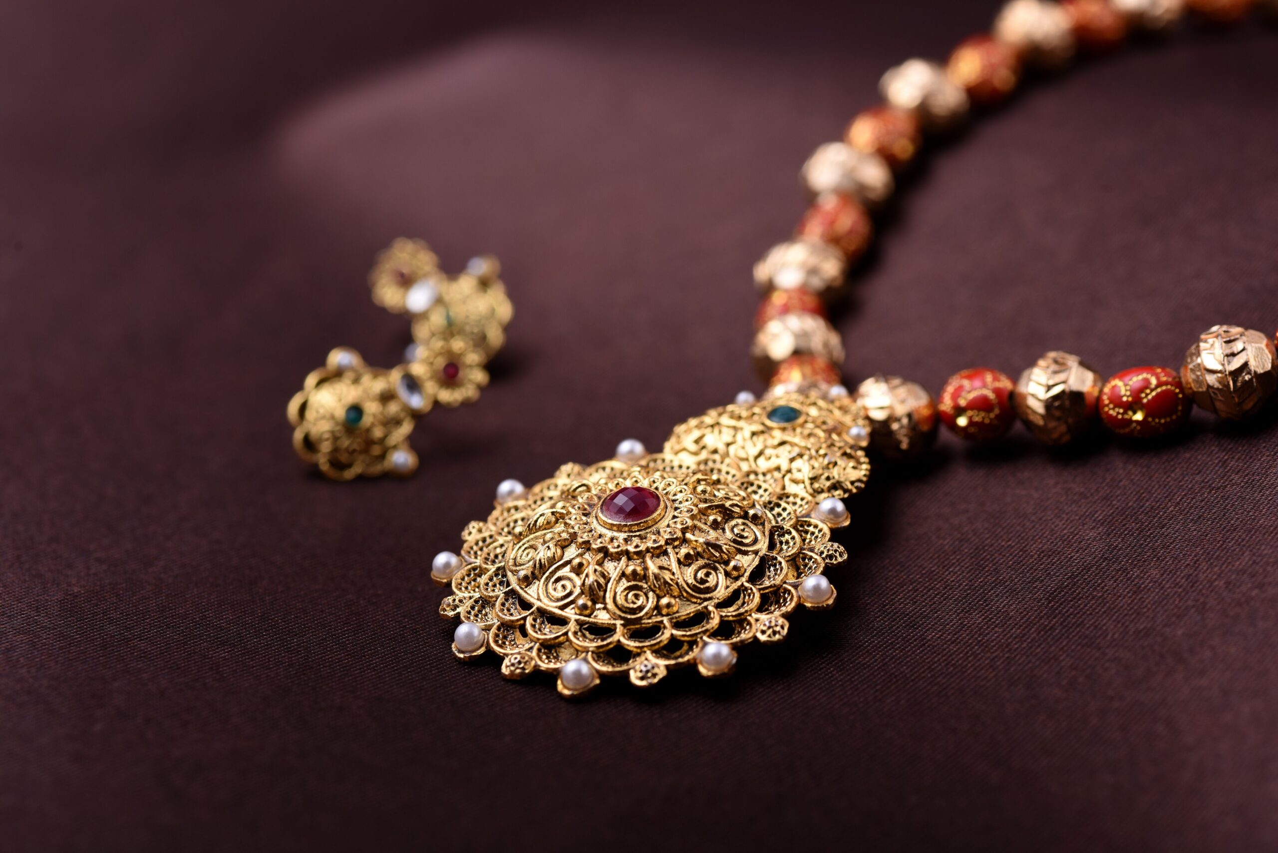 Elegant gold necklace with intricate filigree design, featuring a central red gem and pearls, set against a dark background, exuding a luxurious tone.