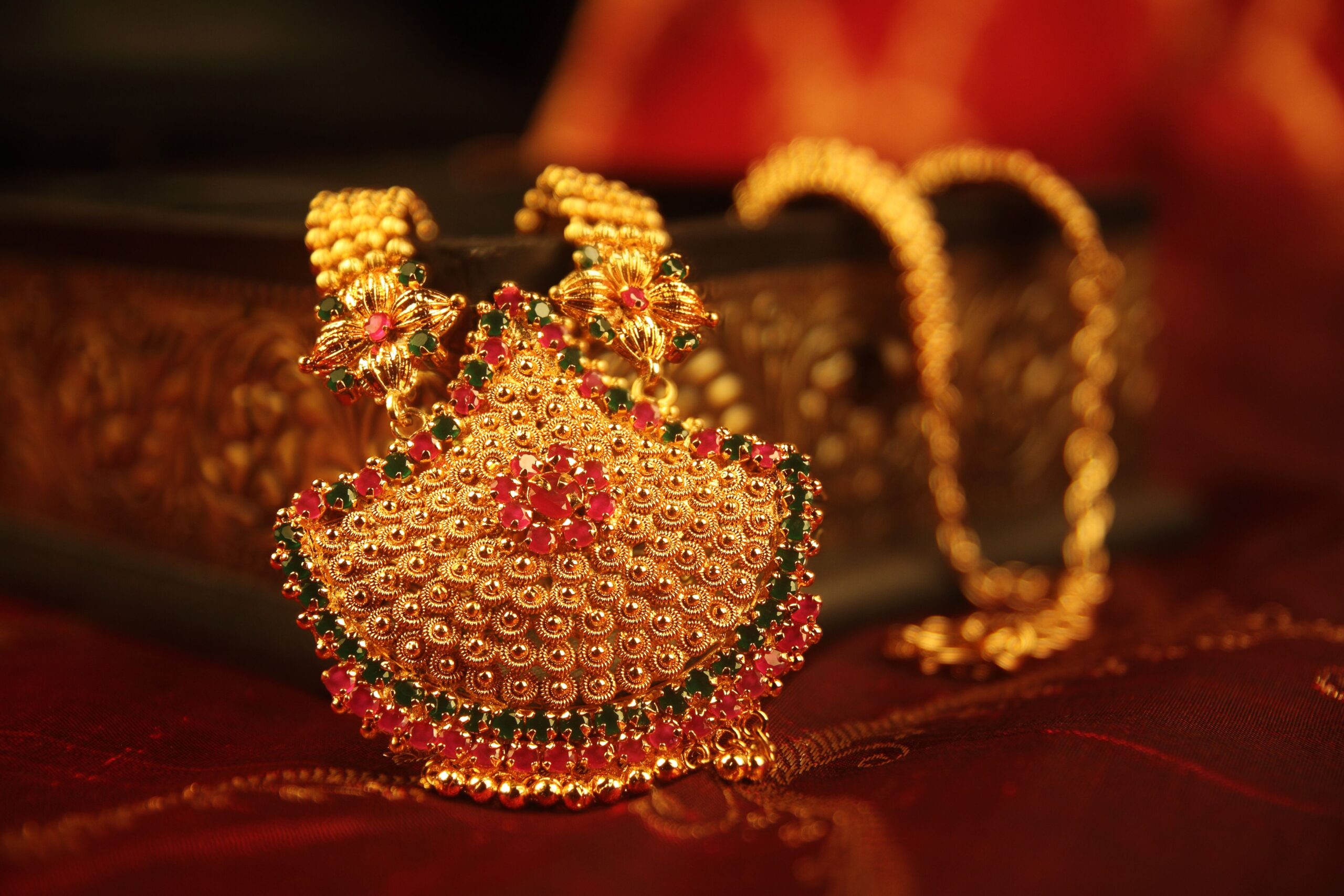 Ornate gold necklace with intricate design, adorned with red and green gemstones, displayed against a richly textured, warm-toned background.