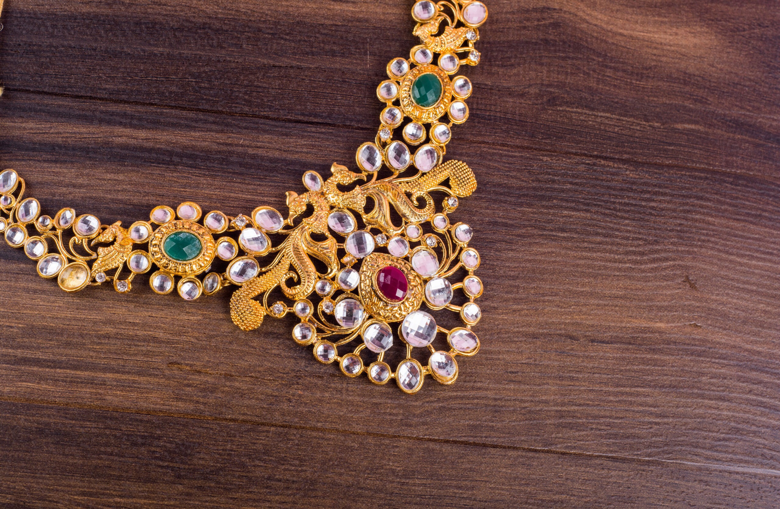 Ornate gold necklace on wooden surface, featuring intricate patterns with green, red, and clear gemstones.