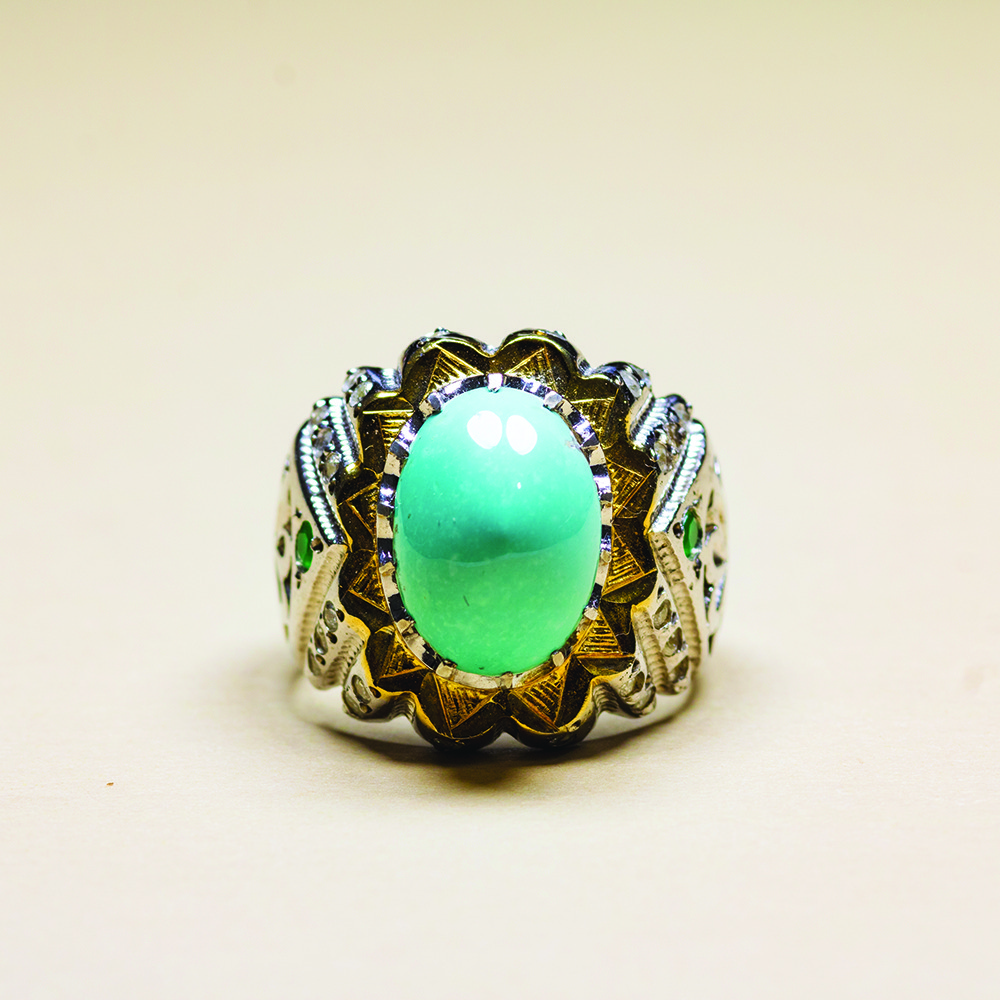 A silver ring with a large oval turquoise stone set in an ornate gold and silver band, featuring intricate patterns and small green gemstones.