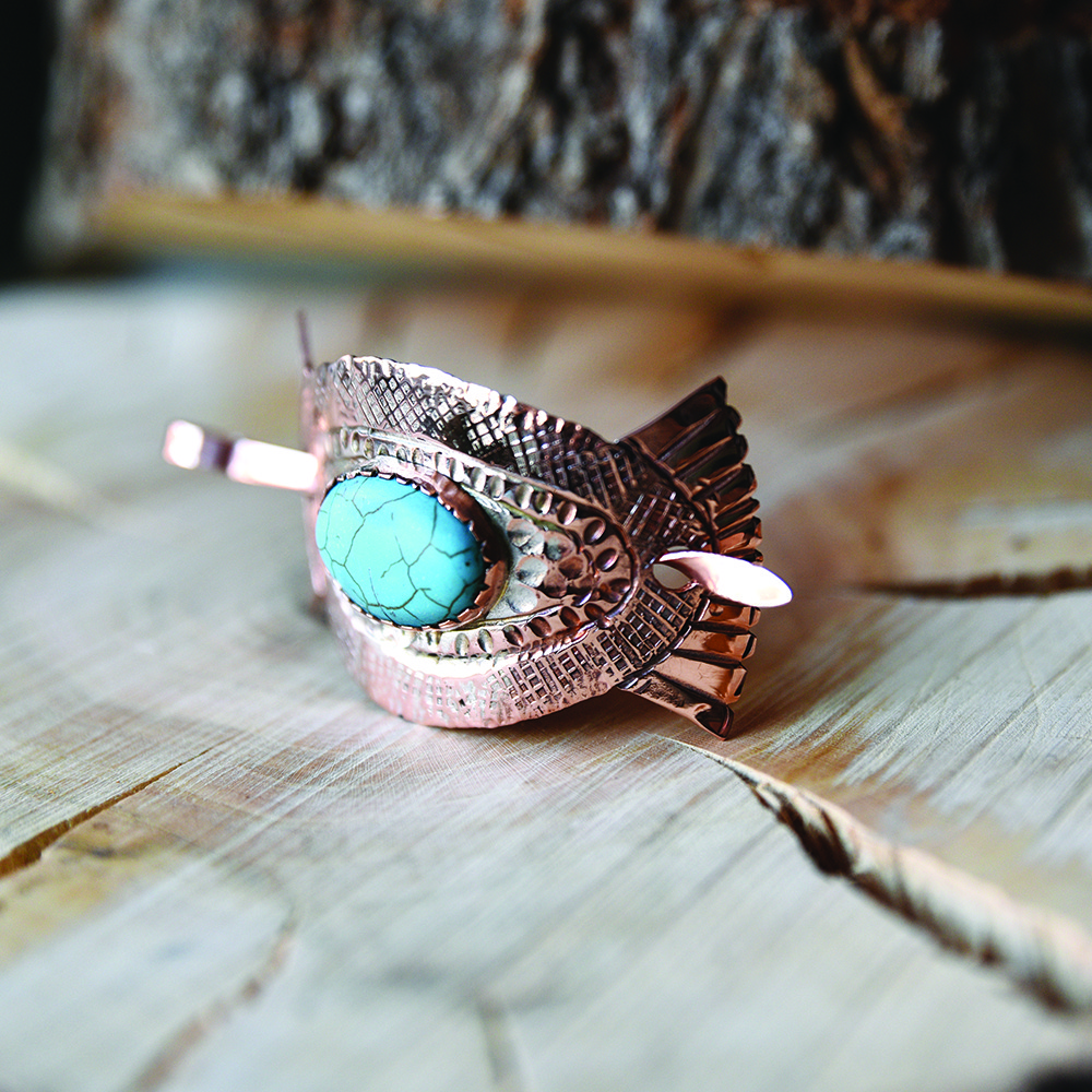 Close-up of an ornate copper bracelet featuring a textured pattern and an oval turquoise stone, resting on a light wooden surface.