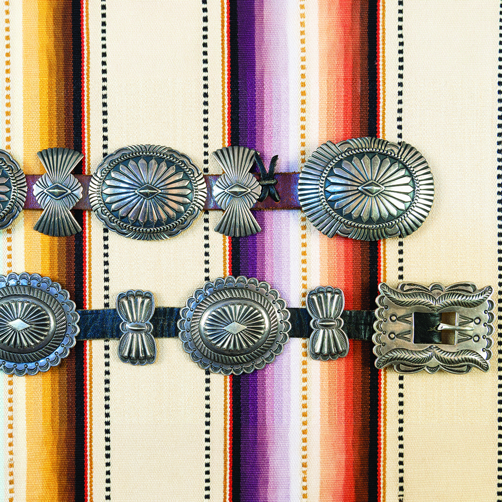 Silver concho belts with intricate designs rest on a colorful, striped textile.