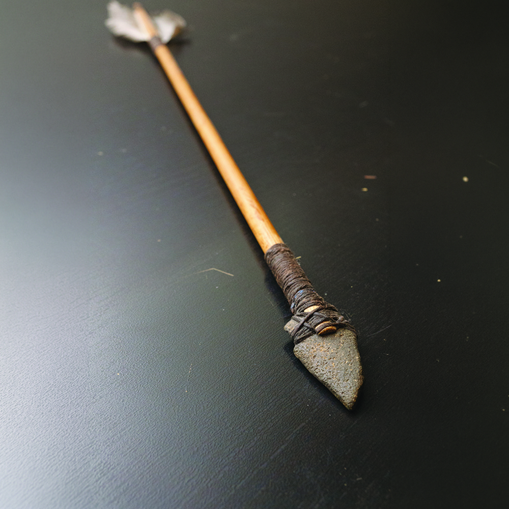 A handmade arrow with a stone tip and feathered end lies on a dark surface.