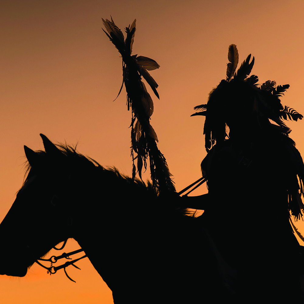 Silhouette of a person in traditional headdress on horseback against an orange sunset sky, conveying a sense of peace and cultural heritage.