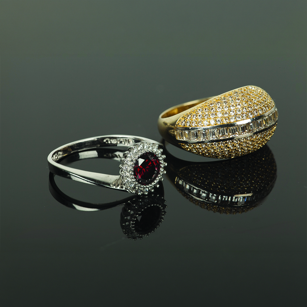 Two elegant rings on a glossy surface: a silver band with a red gemstone surrounded by diamonds, and a gold band encrusted with small diamonds.